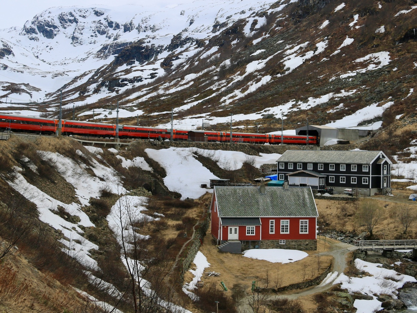 Red passenger train passing snow-covered mountains and wooden houses in Norway